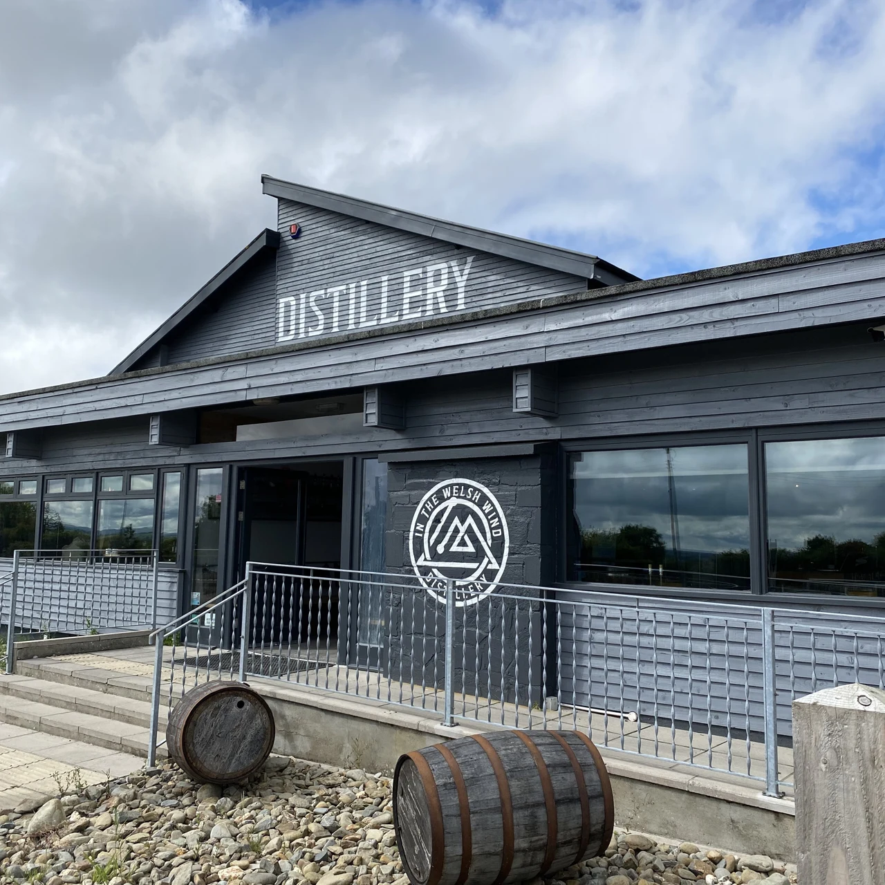 Exterior of In the Welsh Wind distillery, with its entrance, signage and barrels.